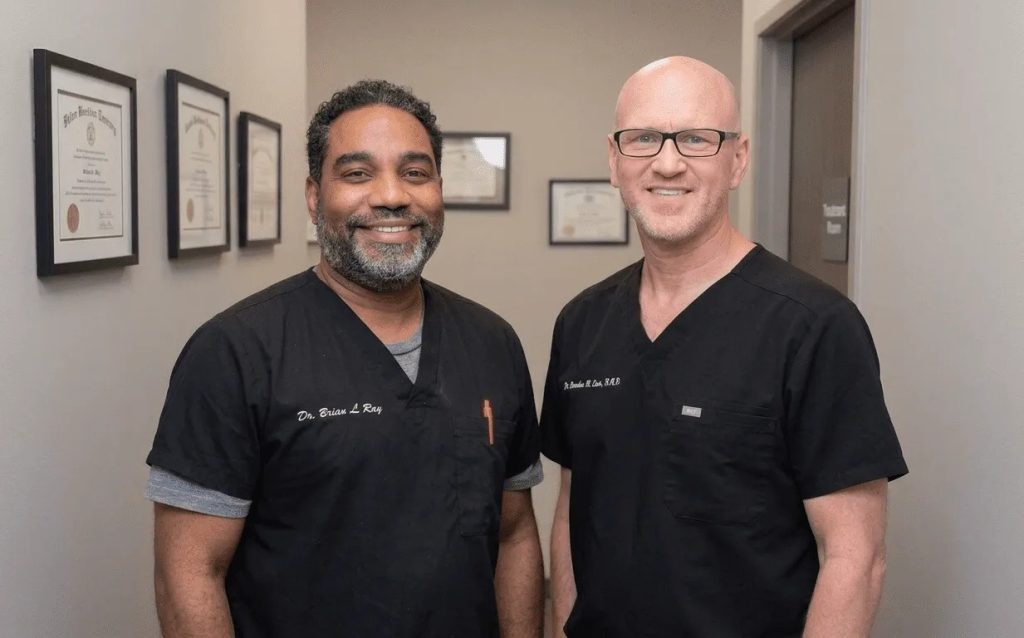 Two smiling dentists in black scrubs standing in front of framed diplomas in the office
